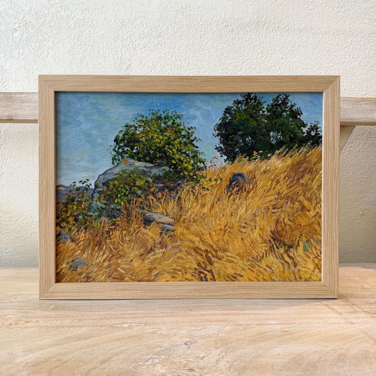Framed painting of a landscape with tall grass and trees on a wooden surface.