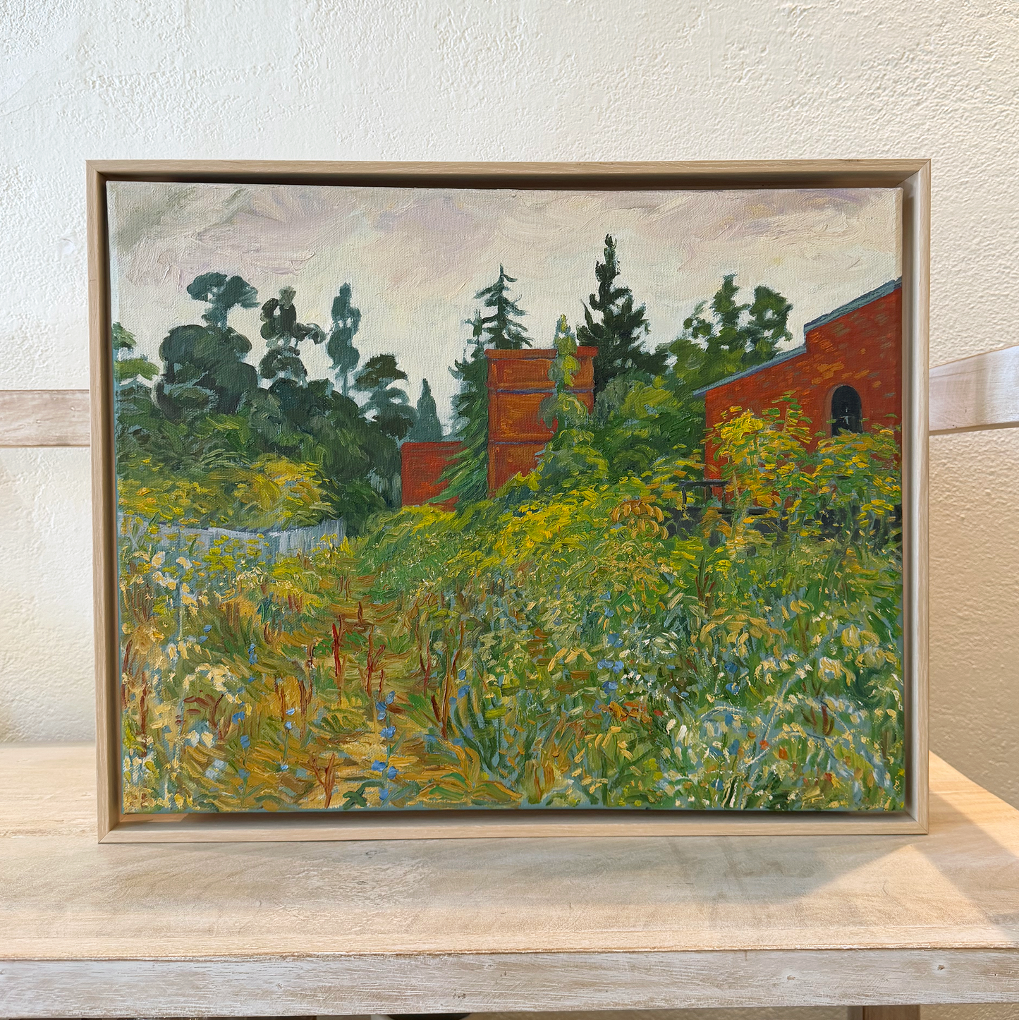 Framed painting of a landscape with trees and buildings on a shelf.