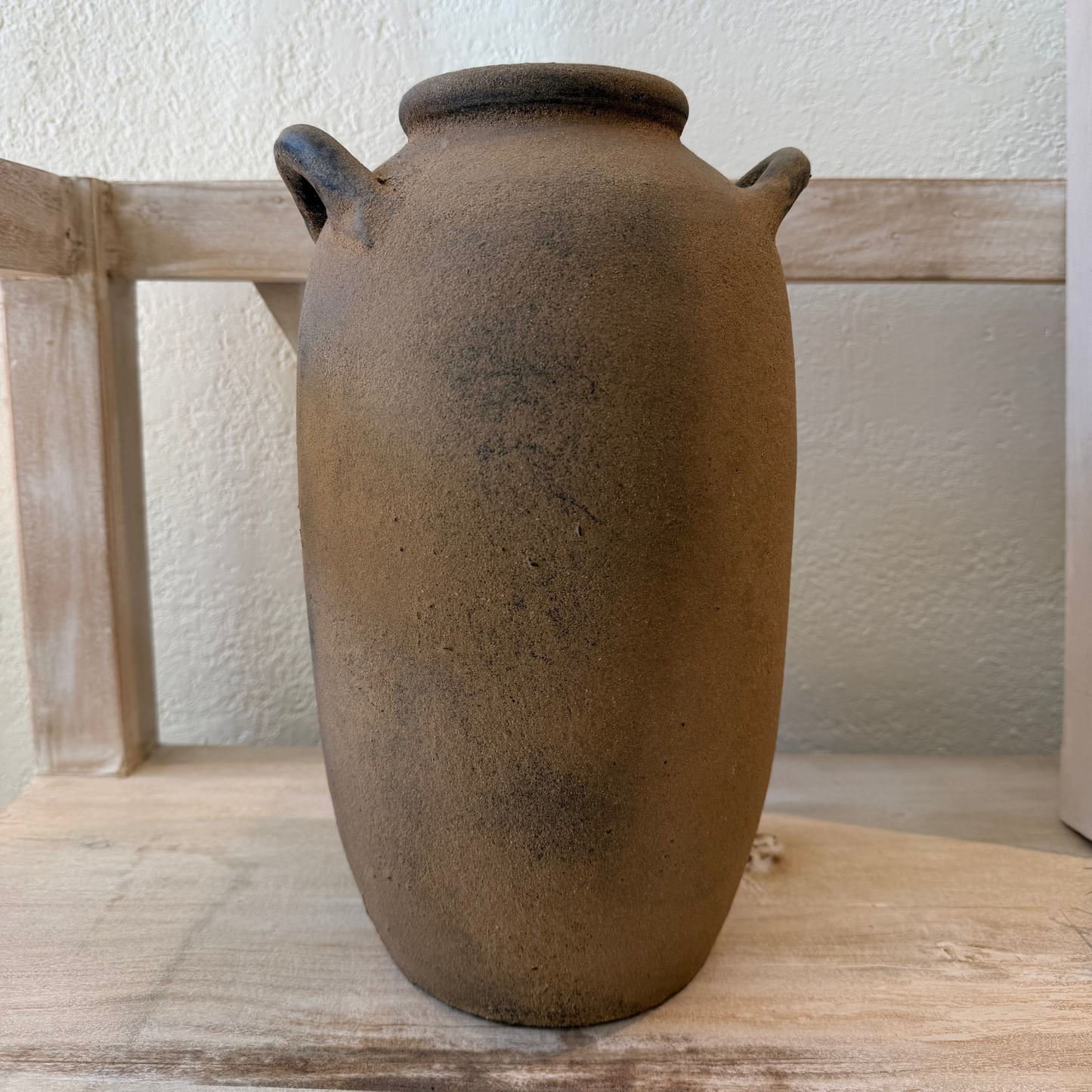 Brown ceramic jar with two handles on a wooden surface against a white wall.