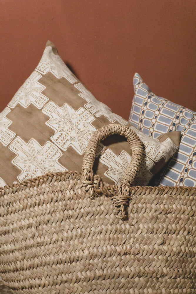 Two block printed throw pillows sit in a handwoven basket in front of a brick colored wall. Each pillow is artisan made and features a unique pattern. One pillow features a large all over print in an earthy brown and the other a smaller scale blue pattern