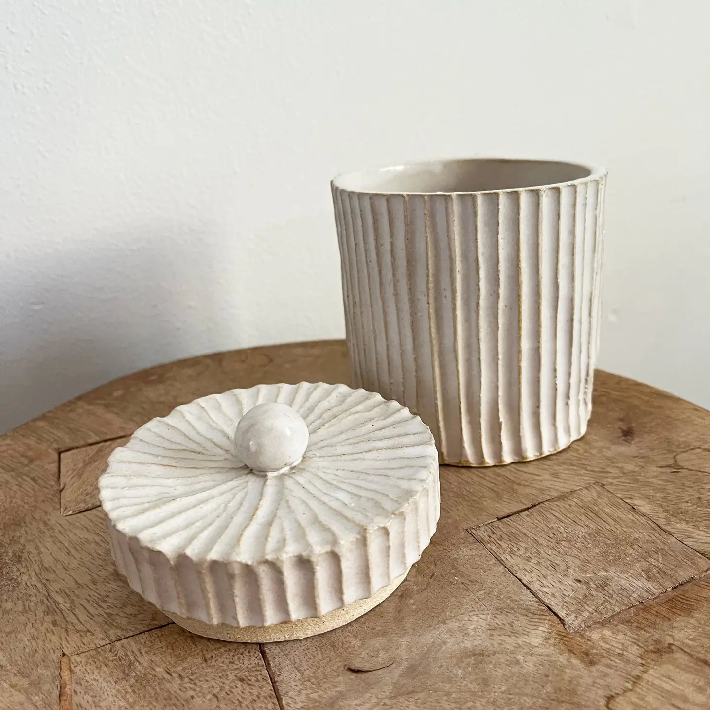 A beige ceramic container with vertical ridges and a round lid, placed on a round  coffee table.
