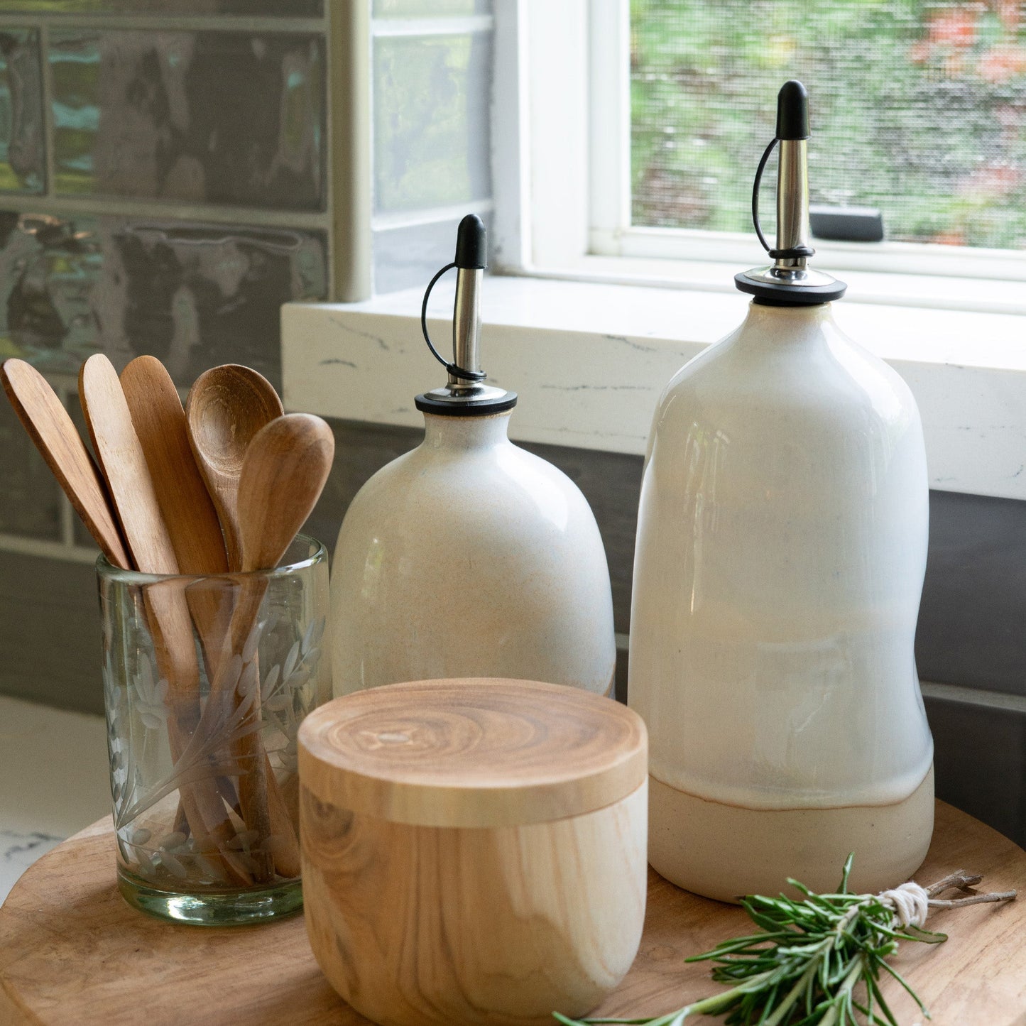 Ceramic olive oil dispenser placed on a chopping board with wooden spoons and a sprig of rosemary  