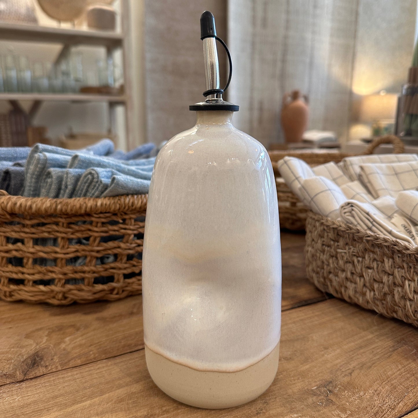 A tall ceramic olive oil dispenser with a natural glaze finish, ergonomic grip, and a black spout, displayed on a wooden surface.