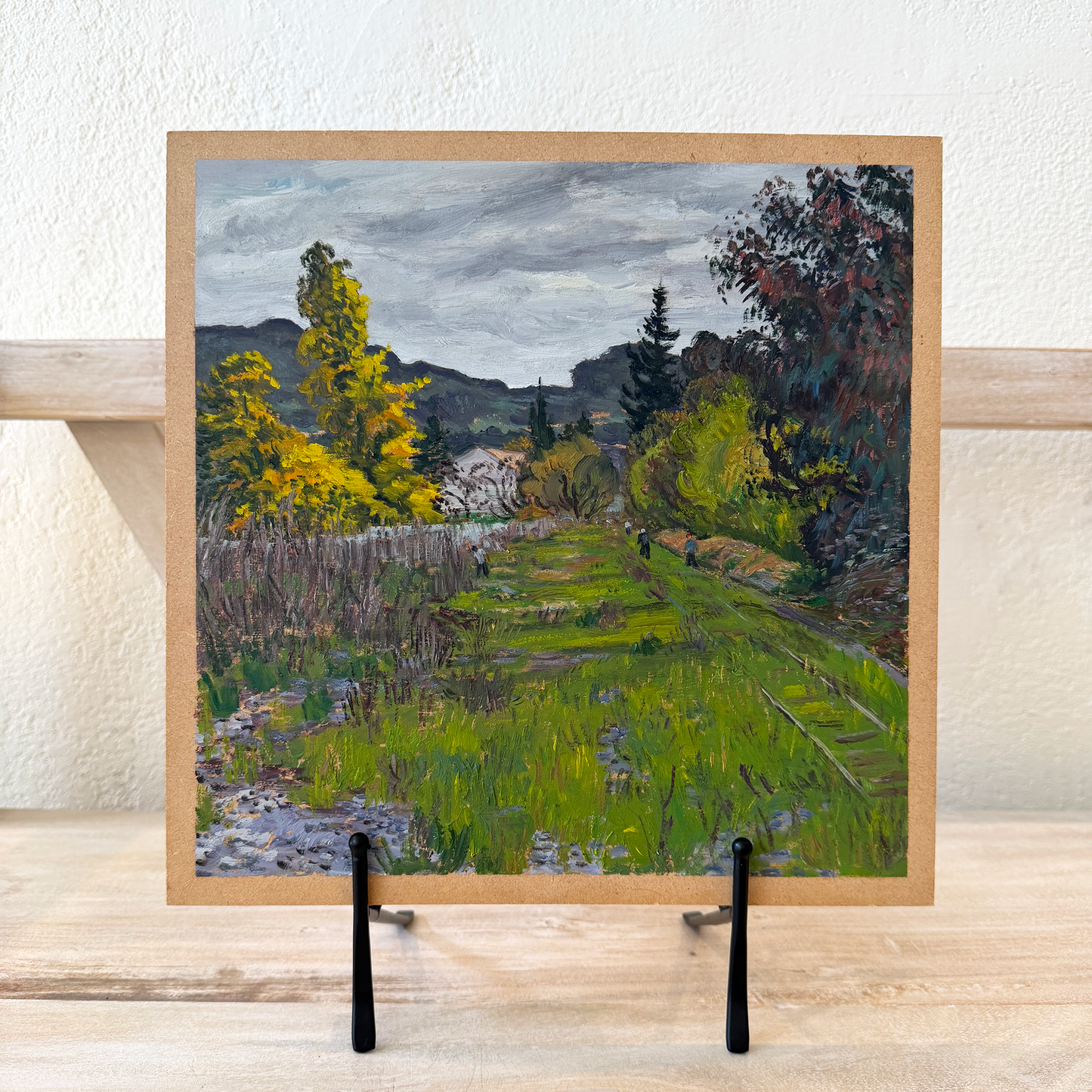 Framed painting of a landscape with trees and a path on a wooden stand.