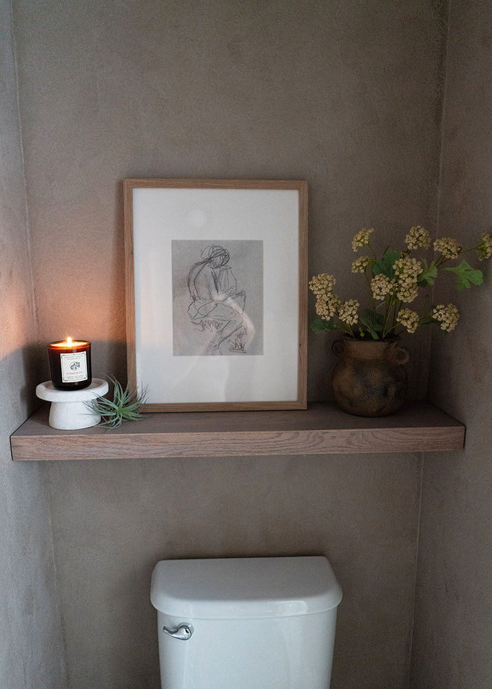 Small shelf with a framed picture, candle, and plant above a toilet in a bathroom.