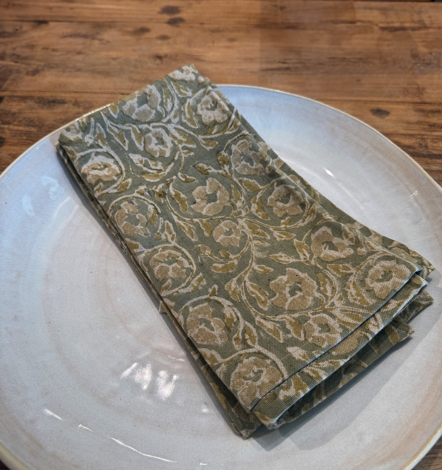 
                  
                    Amara Floral Napkin placed on a hand made ceramic plate showcasing the three tones of green used to make the floral design. The imperfections of block printing are clearly seen.background
                  
                