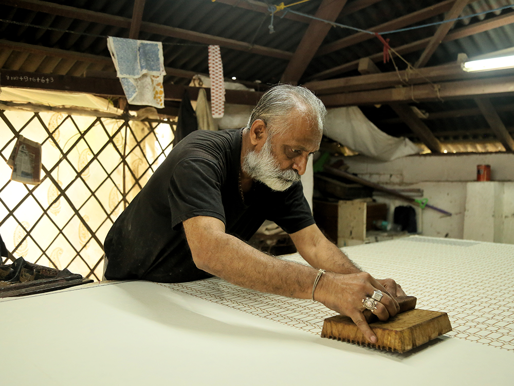 This image is of an artisan block printing fabric on a long table. He is stamping dye onto fabric with a wooden block leaning across the table. 
