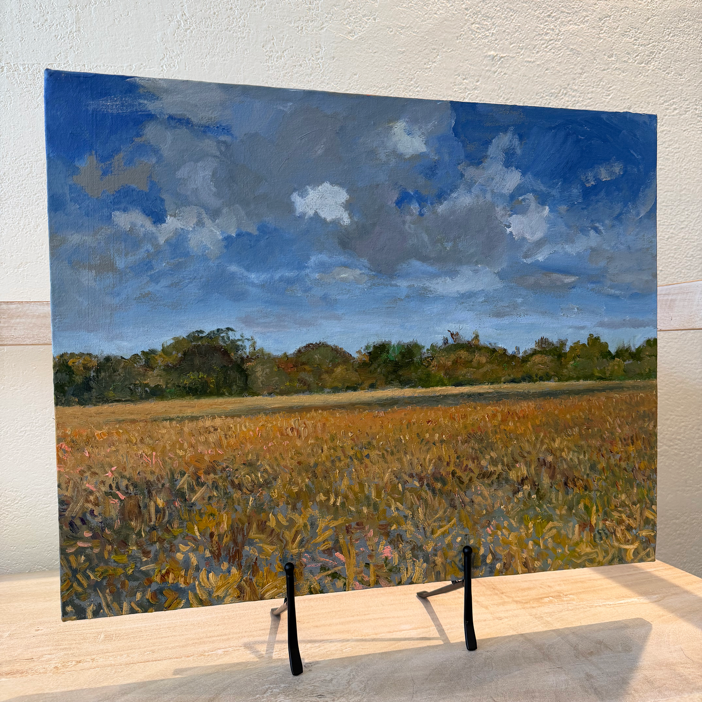 Painting of a field with trees and blue sky on a wooden stand.