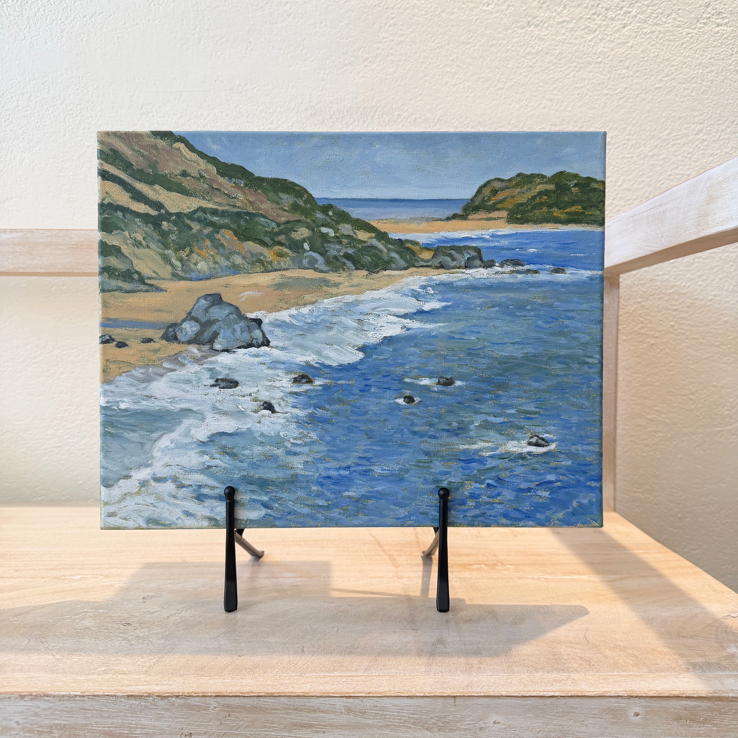 Painting of a coastal scene on a metal easel against a white wall.