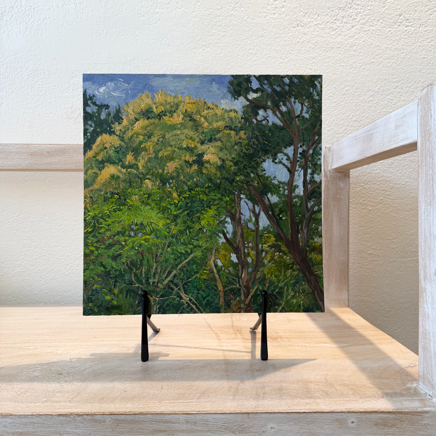 Square painting of a forest scene on a wooden surface with a white brick wall background.
