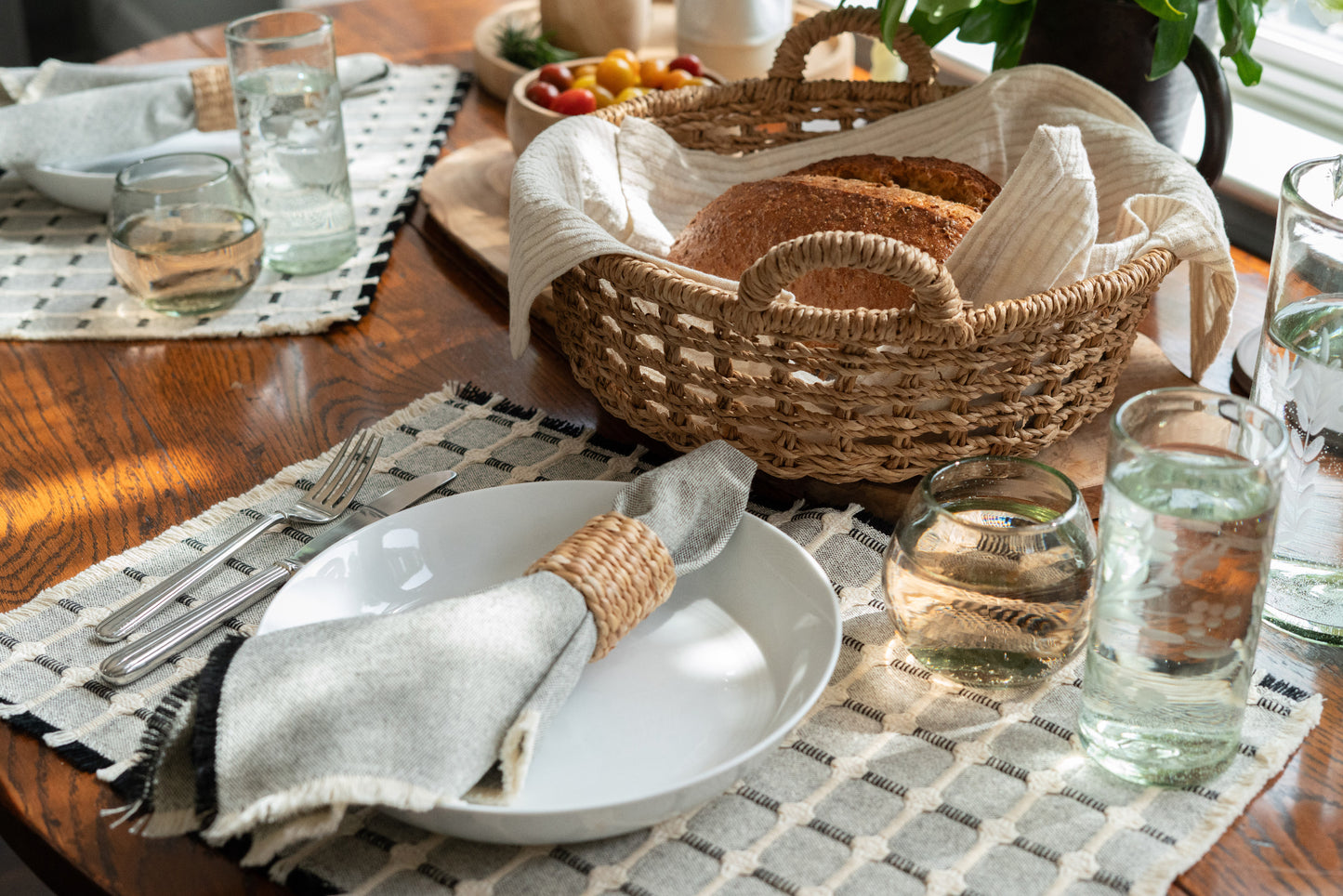 A lite black napkin styled with a reed napkin ring. Placed on a ceramic plate and surrounded by a full table setting including wicker baskets glassware and teak bowls 