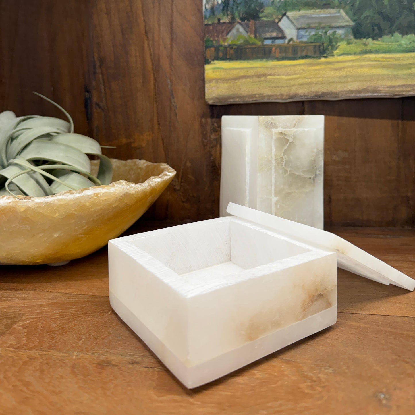 Alabaster Stone Lidded box placed on a dark wood cabinet shelf with two other stone decorative objects background and part of a painting of a landscape in the background