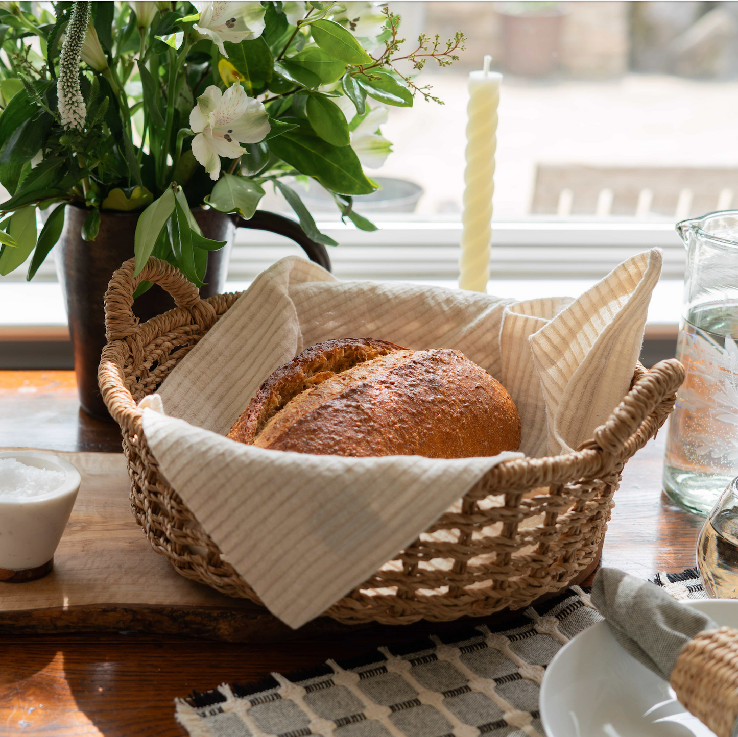 
                  
                    Wicker bread basket with bread on a wooden table with a vase of flowers and candle in the background.
                  
                