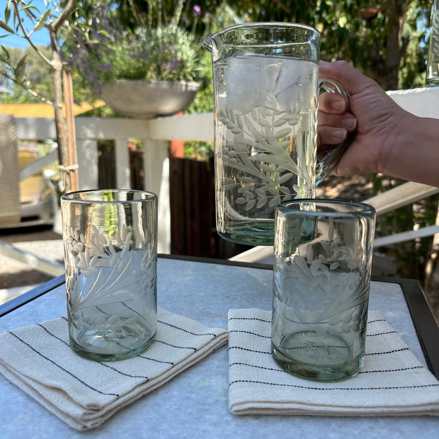 
                  
                    A glass pitcher held in the hand of a designer showcasing the beautiful glass floral etching. A beautiful sunny day in Petaluma
                  
                