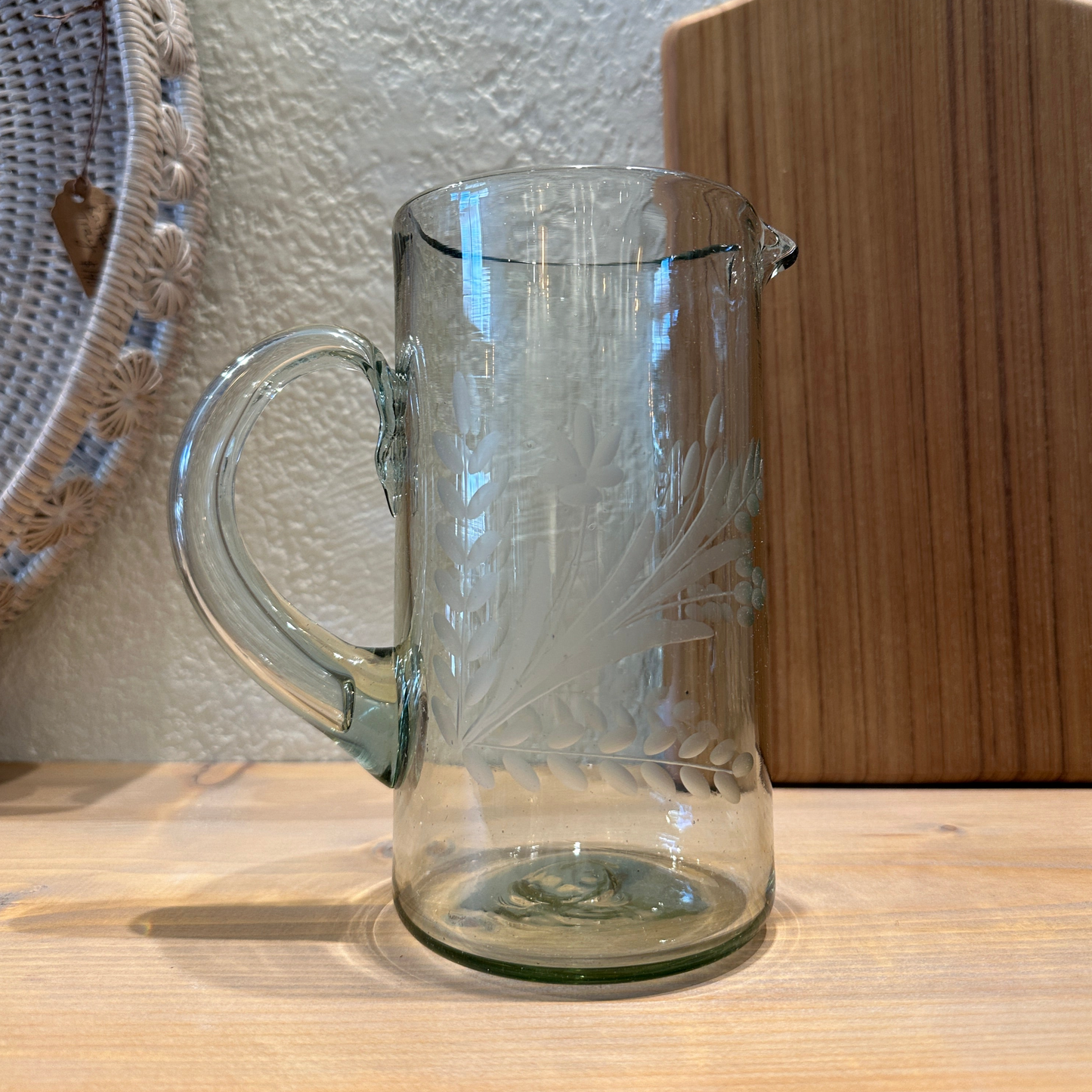 A clear glass pitcher with hand-etched floral designs, displayed against a teak wooden chopping block in the background.