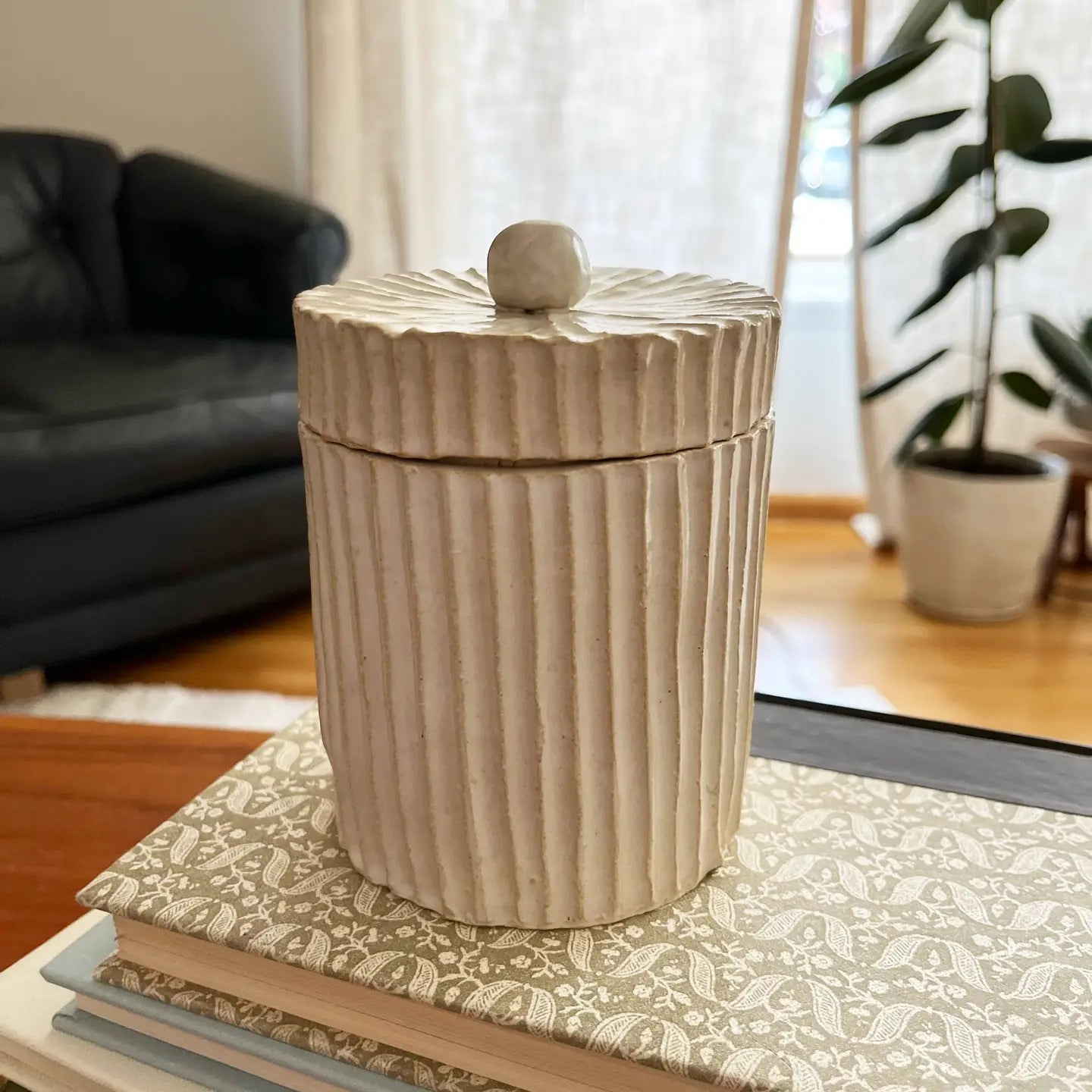 A beige ceramic container with vertical ridges and a round lid, placed on a book on a coffee table.