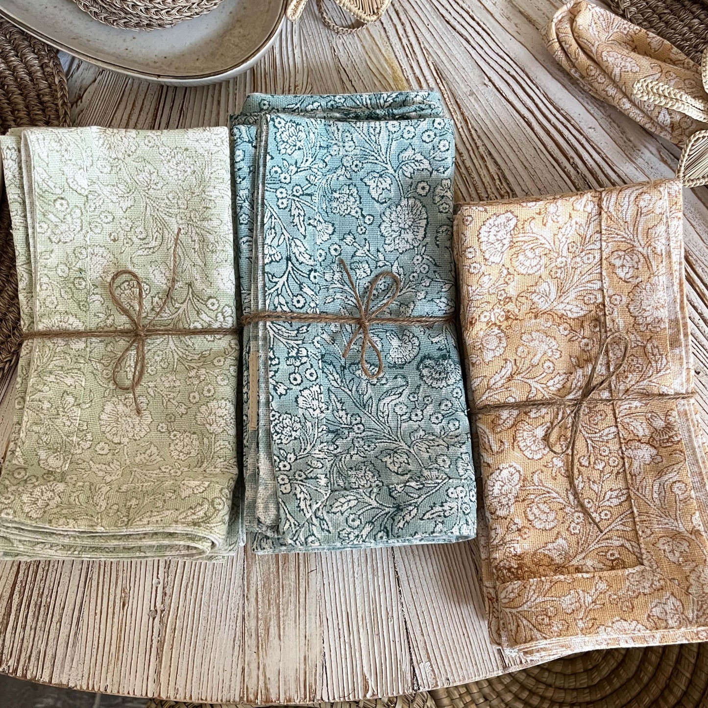 
                  
                    3 Stacks of Floral Napkin's. In three different color ways; Light Green, Blue and Burnt Orange. Displayed in sets of 4, tied together with twin. Placed on a round white washed table in the Petaluma Store
                  
                