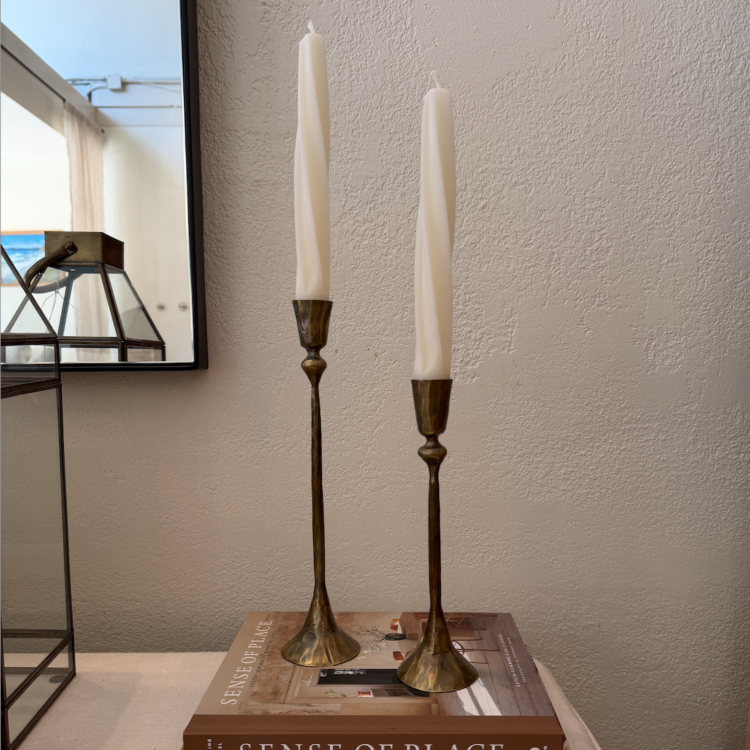 Two tall white taper candles in brass candlestick holders, displayed on a stack of books on a light-colored table against a textured beige wall.