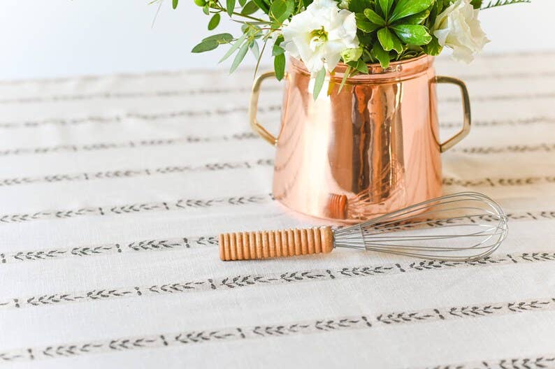 
                  
                    A close up of the wine country style table cloth. Place on top is a cake whisk and a copper vessel with floral arrangement
                  
                