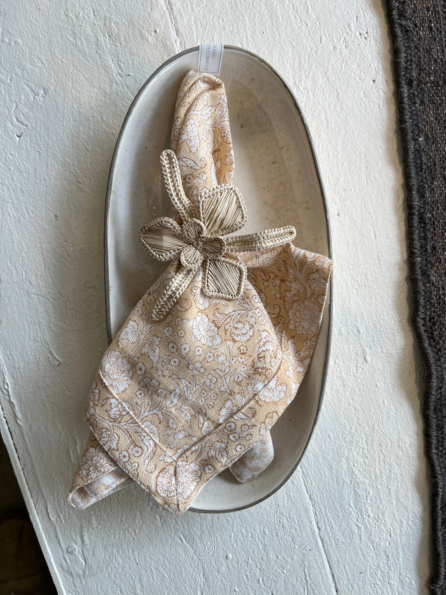 
                  
                    A floral napkin in burnt orange styled in a flower shaped grass napkin ring. This is placed a a hand made porcelain oval serving dish
                  
                