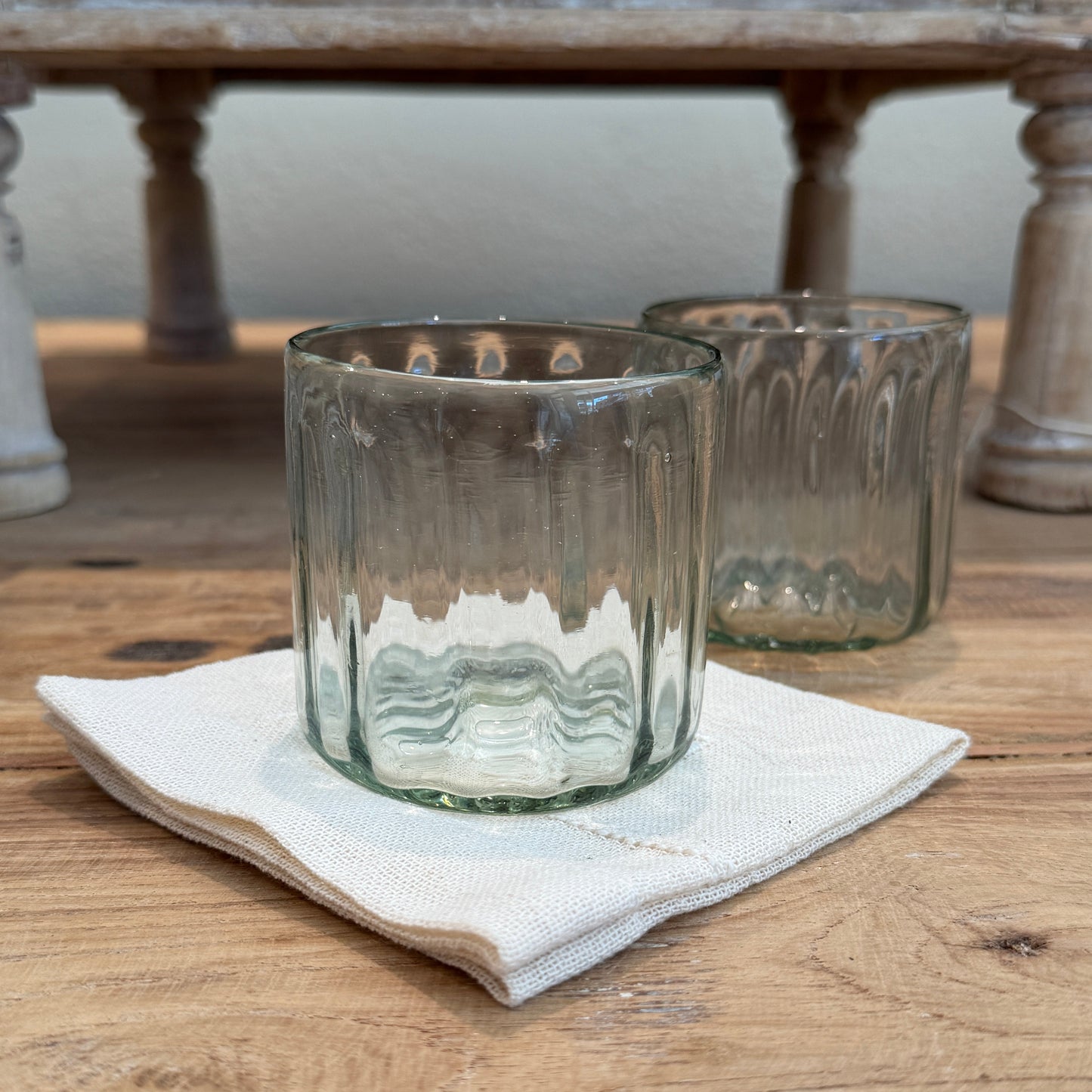 
                  
                    Two ribbed clear drinking glasses or tumblers resting on a folded cream napkin on a wooden table. 
                  
                