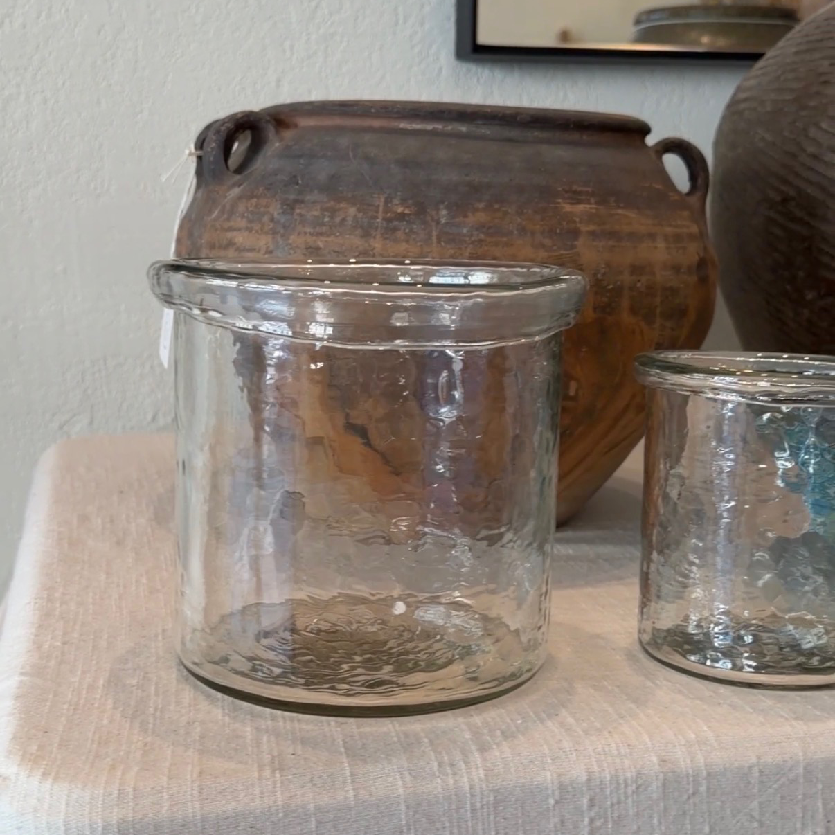 Two clear textured glass canisters or candle holders displayed on a light beige fabric-covered surface, with a rustic brown ceramic pot in the background.
