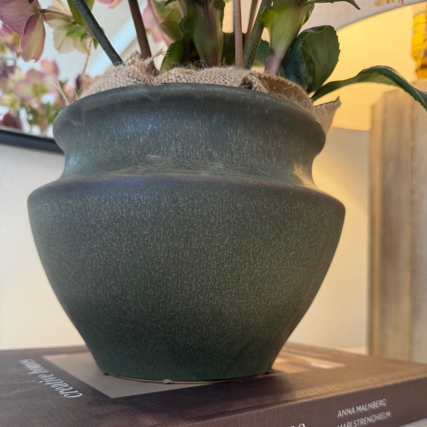 
                  
                    Dark green ceramic vase on a book titled 'creative homes' with flowers in the background.
                  
                