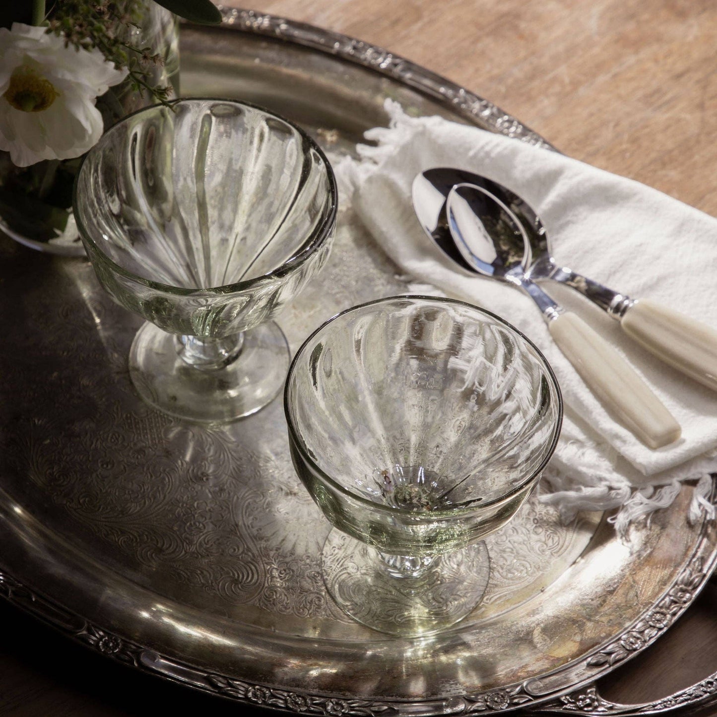 
                  
                    Two glass dessert bowls on a silver tray with white flowers and cutlery.
                  
                