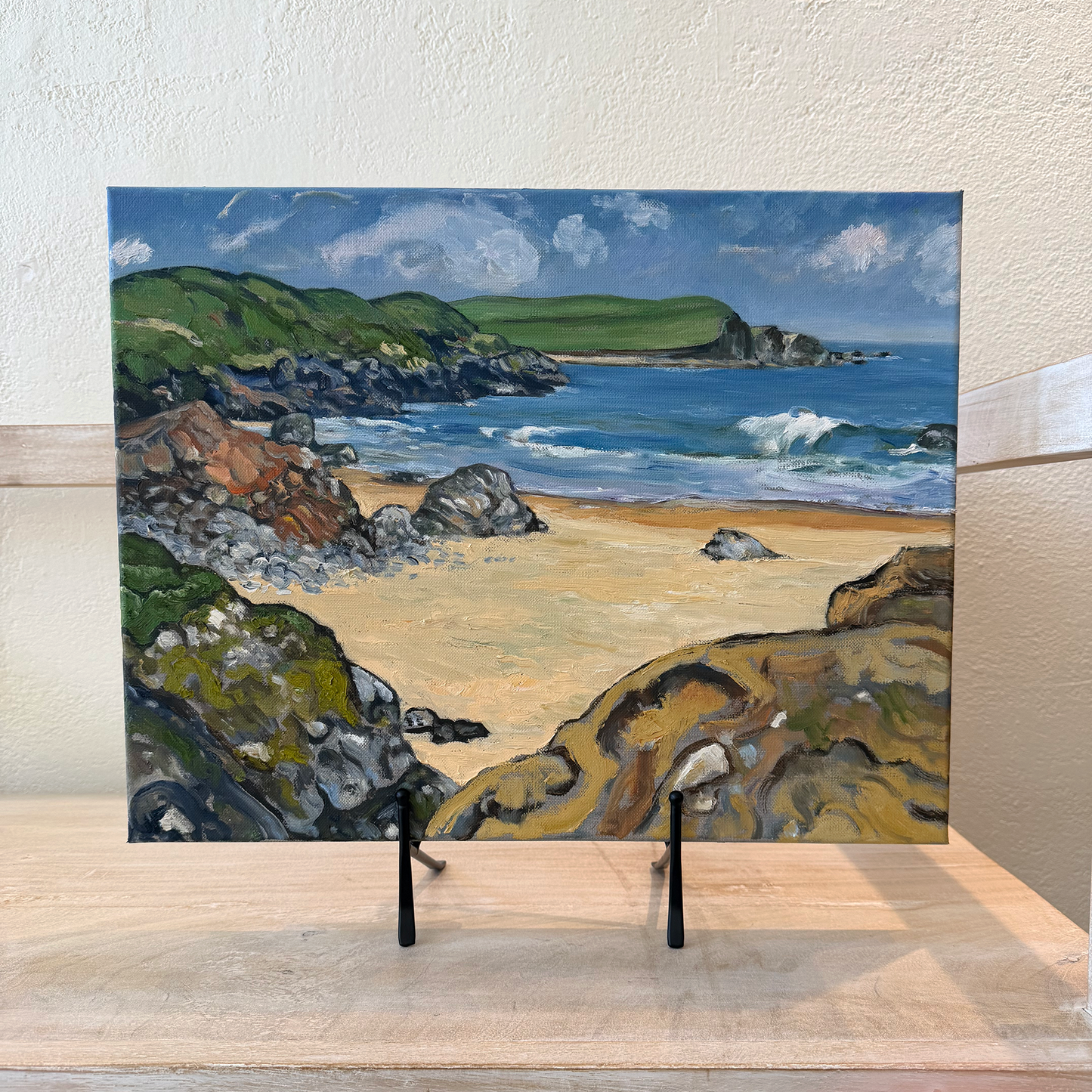 Painting of a coastal scene with rocks, beach, and ocean on a wooden surface.
